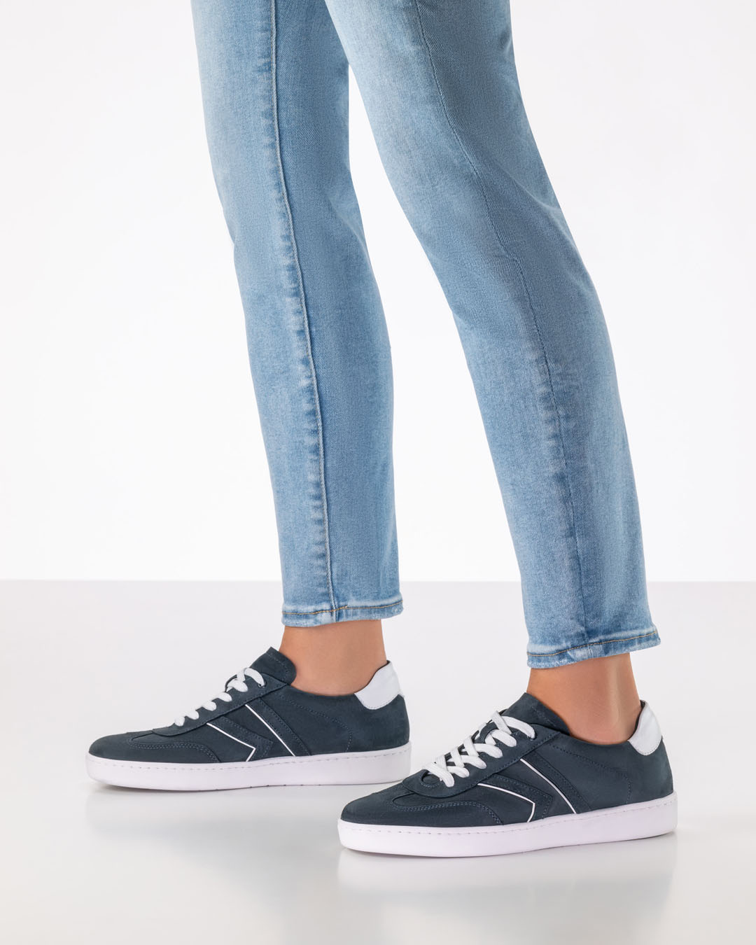 The model is standing to one side, wearing light-coloured, slim-fit jeans and dark blue suede trainers with white soles