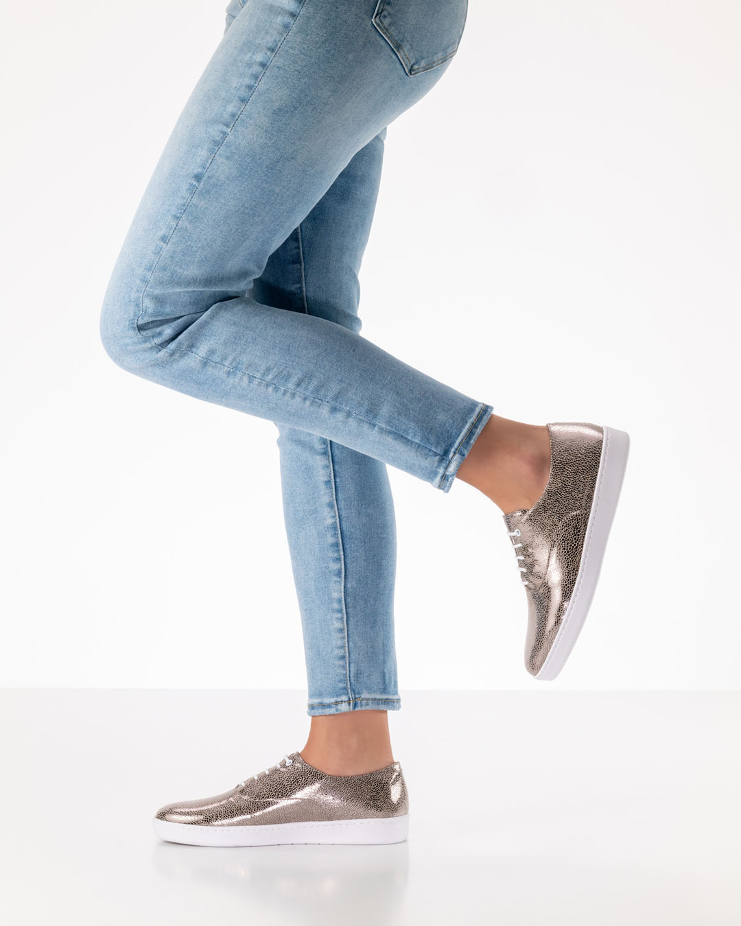 The legs of a dance shoe model wearing light-coloured jeans and platinum-coloured lace-up dance shoes with white PU soles