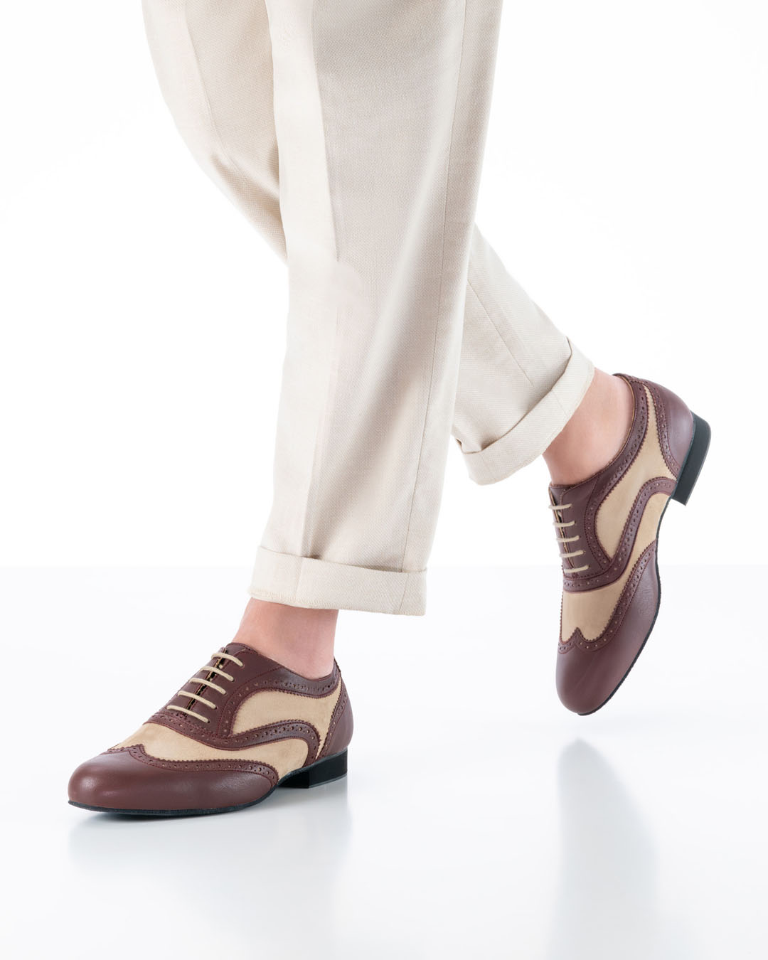 A male model in cream-coloured trousers walks towards the camera wearing caramel and burgundy dancing shoes with a brogue pattern