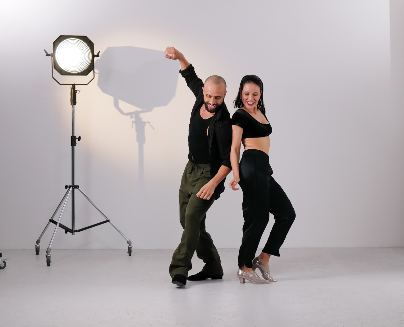 Bachata-3 Dynamic bachata moves by a young dance couple in casual dark outfits wearing silver and black dance shoes by Werner Kern in front of a white studio wall with spotlights in the background.