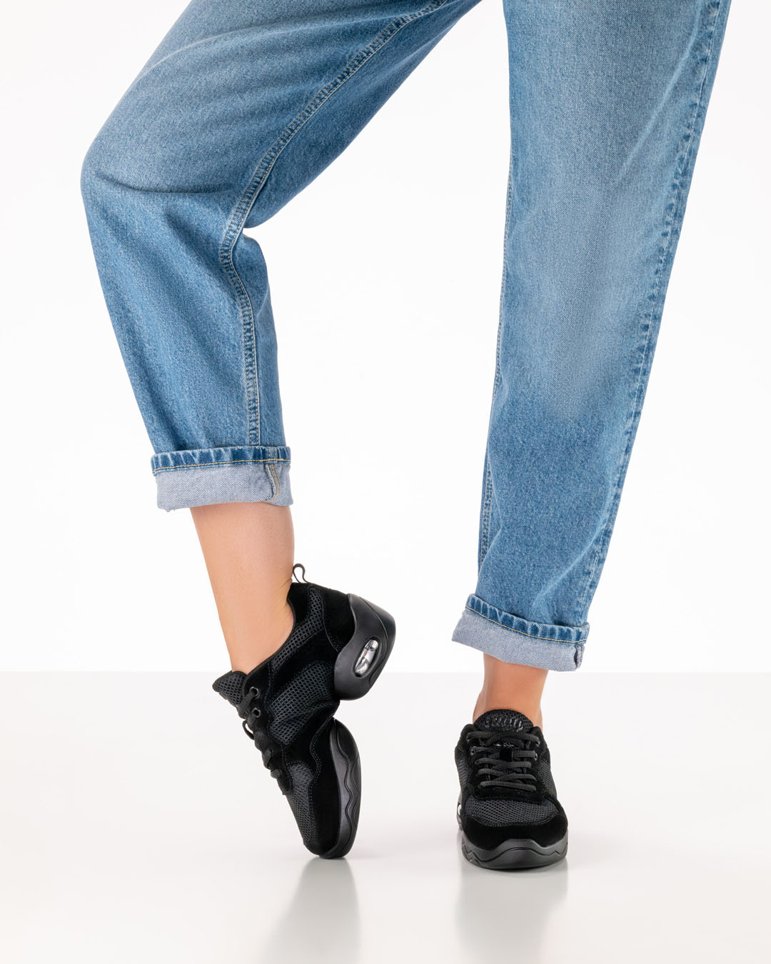 A model in jeans is wearing black dance trainers with a split sole and is bending her right foot over the instep to demonstrate the shoe’s flexibility
